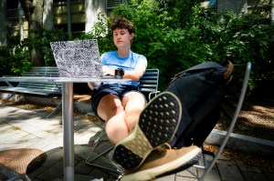 A student relaxes outside at Northeastern University, working on a laptop with coffee in hand and their feet propped up on a nearby chair.