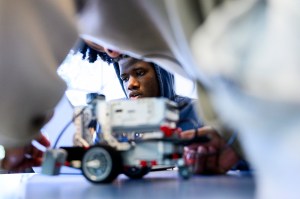 A student leans in to adjust a small wheeled robot on a classroom table, framed by the blurred arms of classmates.