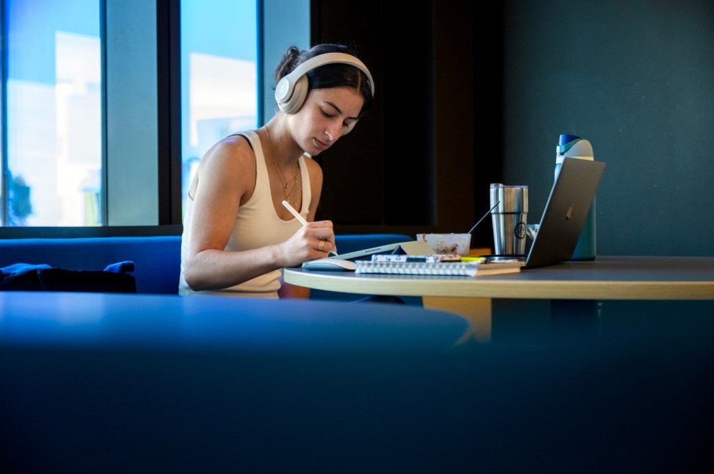 A person wearing headphones studies at a table with an iPad and a laptop.