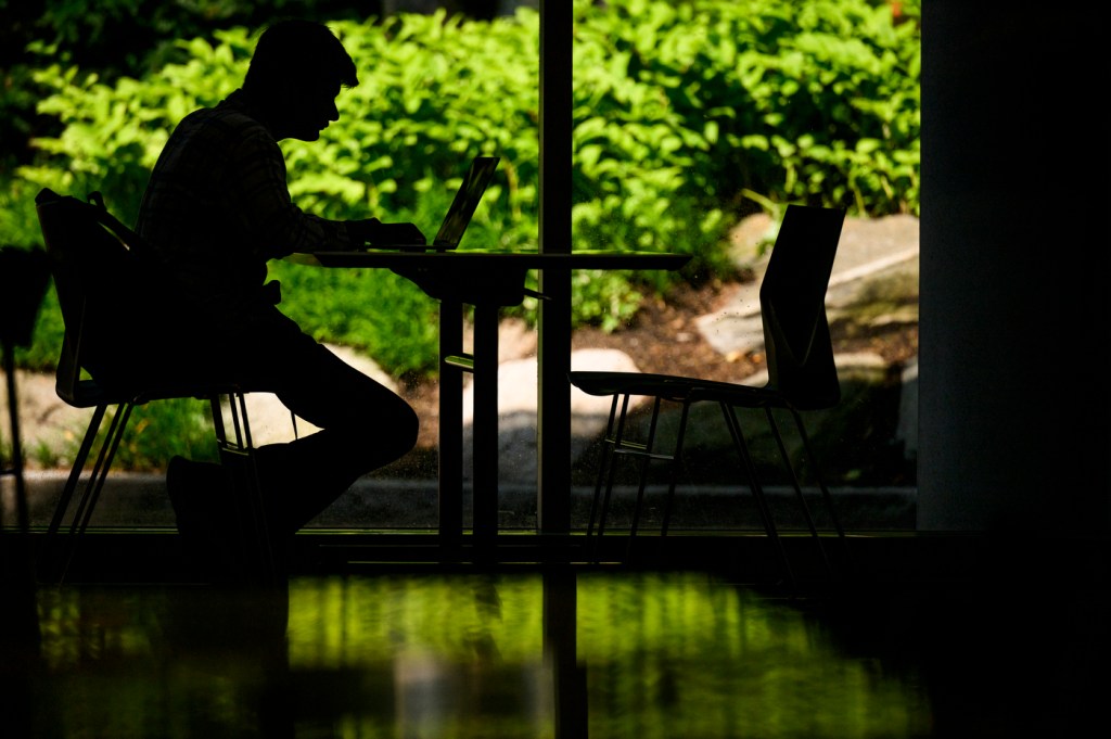 The silhouette of a student sitting in front of windows working on a project. In the background, there is green foliage outside of the window.