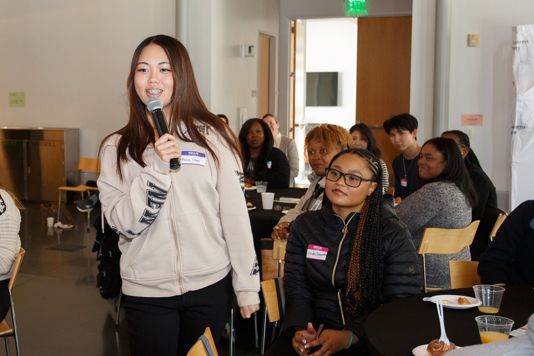 A student presenter in a beige sweater speaking in a microphone to seated attendees wearing name tags