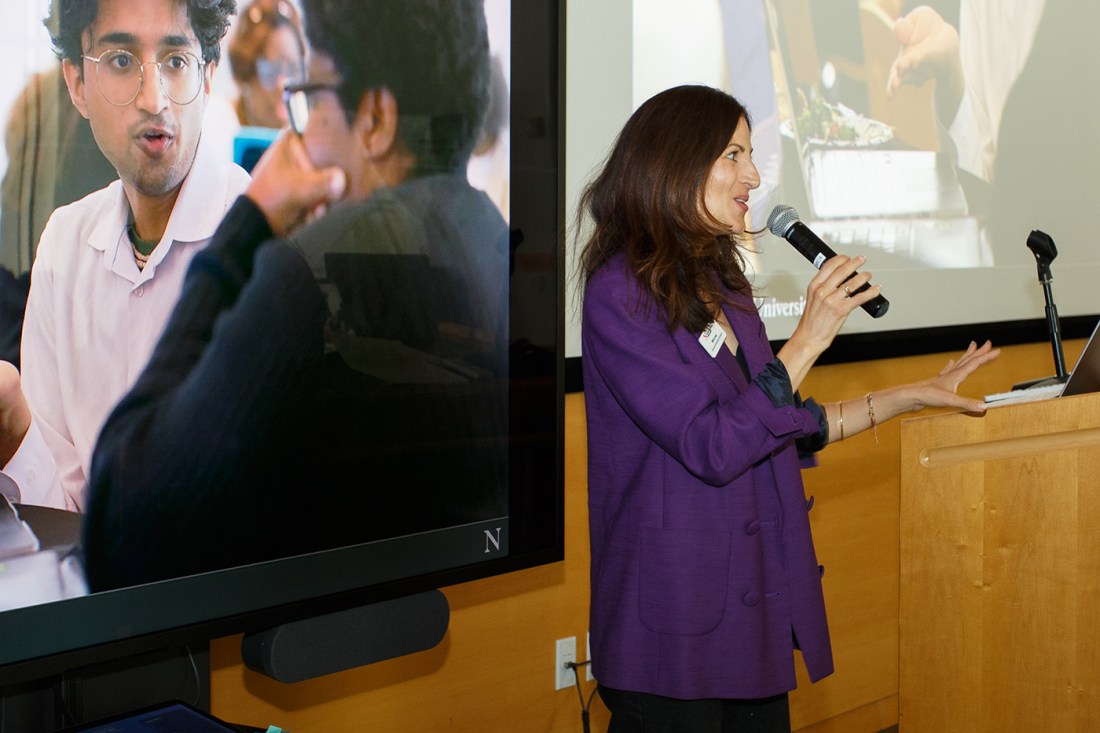 A woman in a purple blazer speaking at a podium with a microphone, in front of a screen showing an image of someone in a white lab coat speaking with another person.