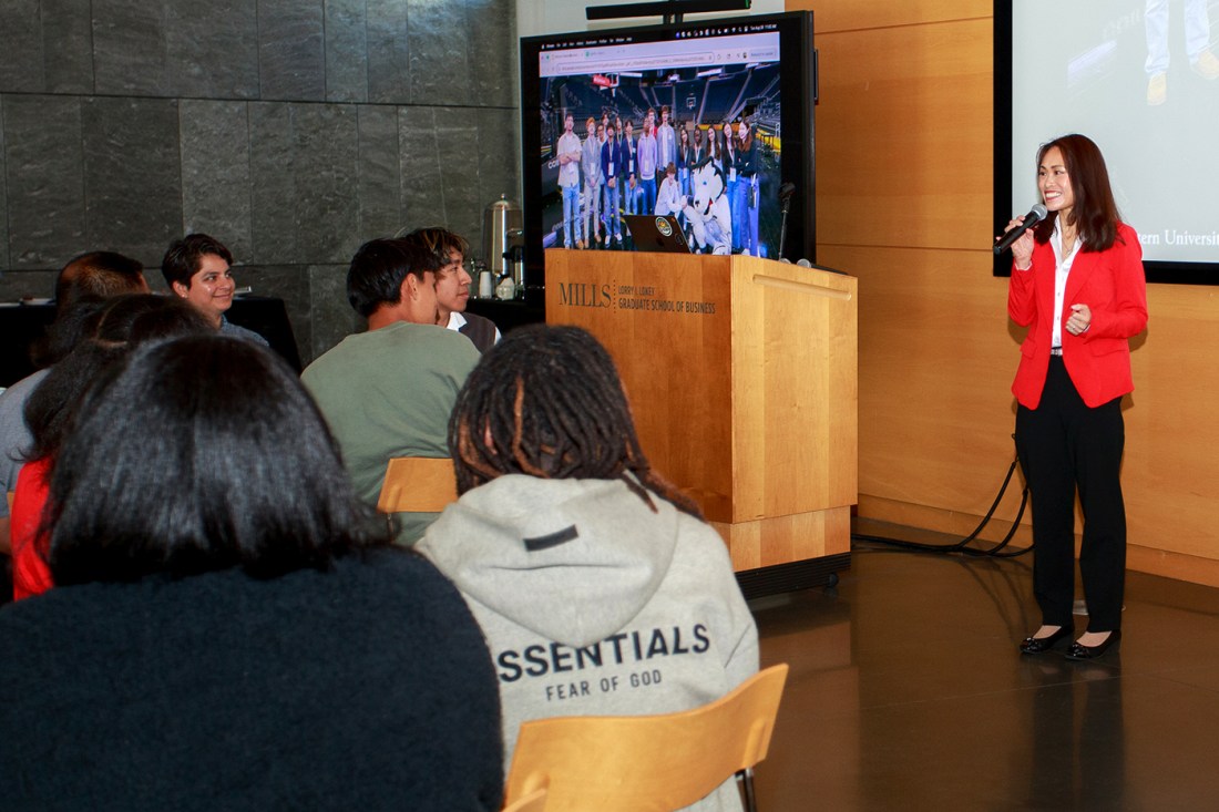 A presenter in a red blazer at a podium with a screen displaying a group photo behind her, with attendees seated at tables