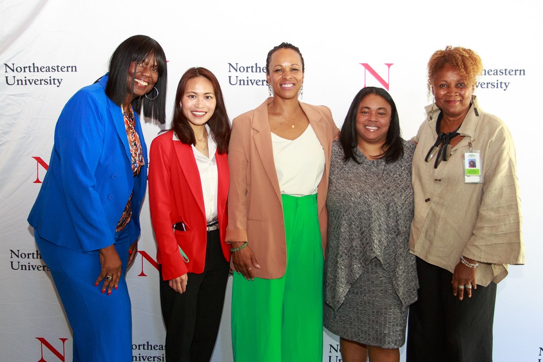 Five women posing together in professional attire including some wearing colorful blazers in blue, red, tan, and green against a Northeastern University backdrop.