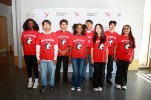 Eight students wearing matching red Northeastern University t-shirts with the Husky mascot logo stand together in two rows for a group photo. They are positioned in front of a white backdrop featuring repeated Northeastern University branding.