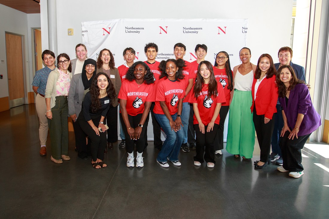 A large group photo of approximately 20 people, with some wearing red Northeastern t-shirts with the Husky logo, standing together in front of a Northeastern University branded backdrop.