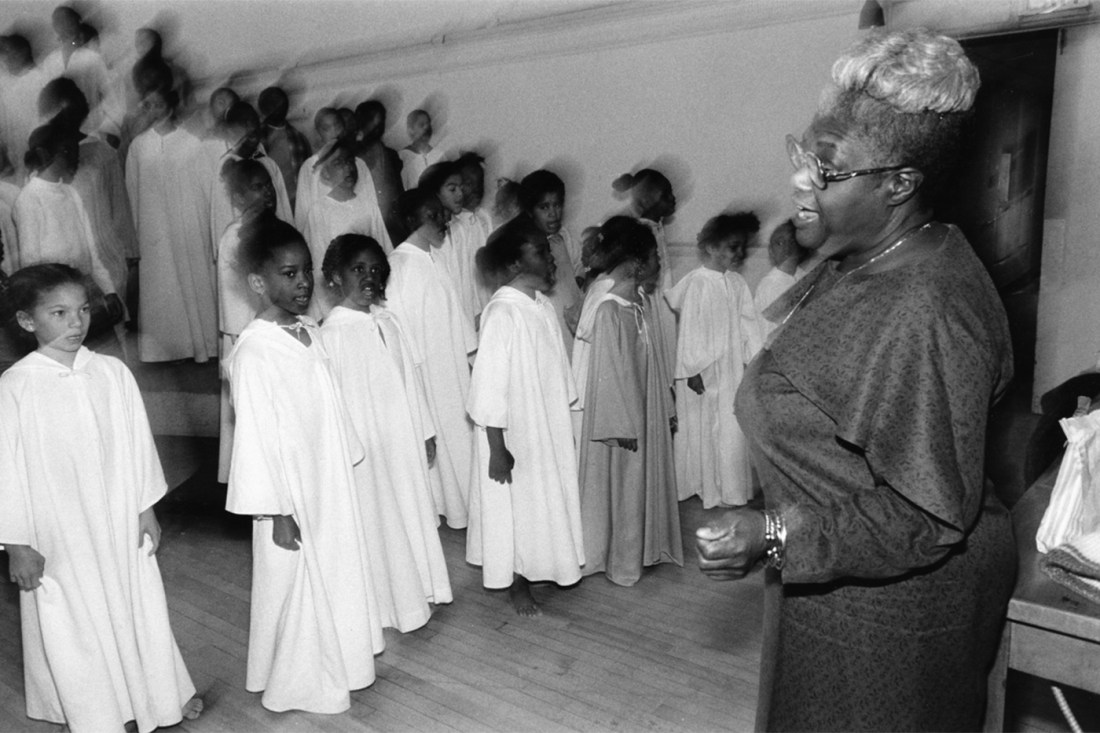 A black and white photo of Elma Lewis speaking to a chorus of children in white robes.