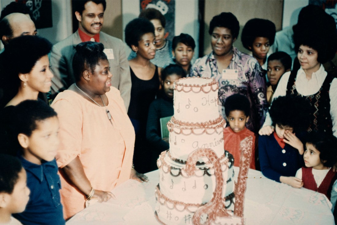 Elma Lewis and many others smiling and surrounding a table with a large tiered cake that has the number 21 prominently in front.