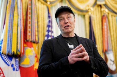 Elon Musk speaks with hands clasped, wearing a black jacket and hat, standing in front of military flags and gold curtains.