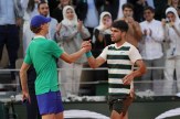 Jannik Sinner, wearing a green shirt and blue hat, shakes the hand of Carlos Alcaraz, wearing a striped shirt, at a tennis net.