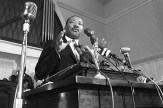 A black and white historical photo of Rev. Martin Luther King Jr. speaking at a podium in front of several microphones.
