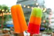 Close-up of two colorful ice pops held outdoors, with blurred summer lights and buildings in the background.