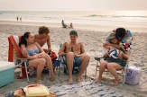 A group of teens relax on the beach in lawn chairs, laughing and chatting as the sun sets over the ocean.