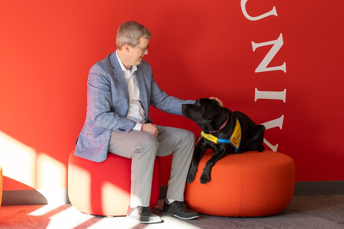 Dean Thurman wearing a suit sitting on a red chair against a red wall petting a black dog wearing a yellow and blue service vest.