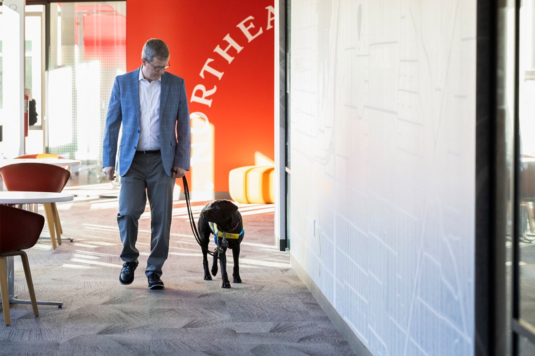 Dean Thurman wears a blue suit walks through a modern office corridor along a black dog wearing a yellow and blue service vest. The hallway has white walls and red accent elemnts visible in the background.