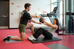 A personal trainer kneels on one knee to pass a weighted ball to a woman doing sit ups on a mat on the floor.