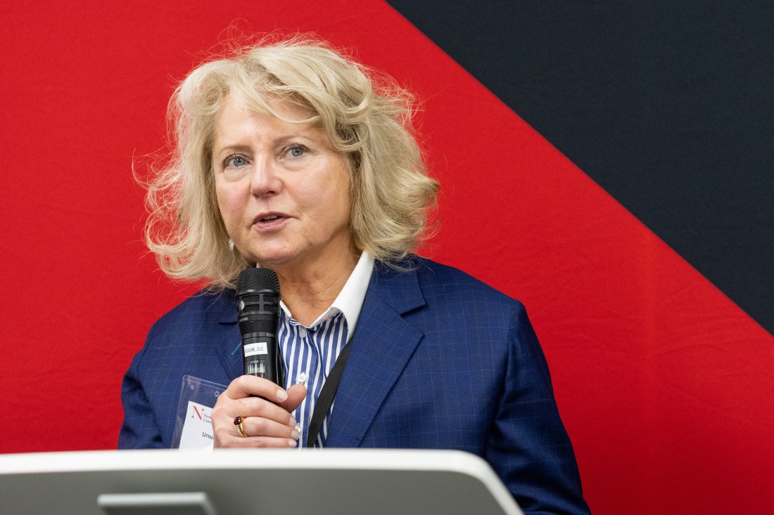 Ursula Smartt depicted in a suit blazer speaking into a microphone against a red backdrop.
