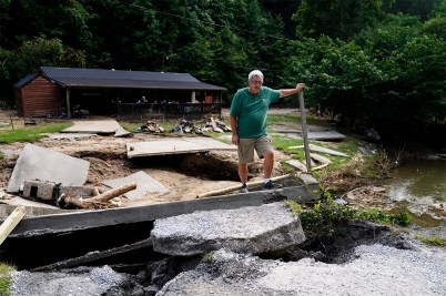 A man wearing a green t-shirt and khaki colored shorts standing amongst the rubble of a home destroyed by flooding.