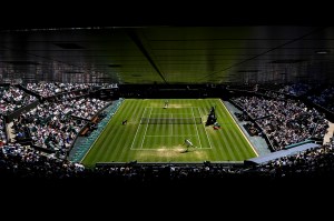 Aerial view of a tennis court during a match, showing the grass court from above with two players positioned on opposite sides. The court is surrounded by packed stadium seats filled with spectators.