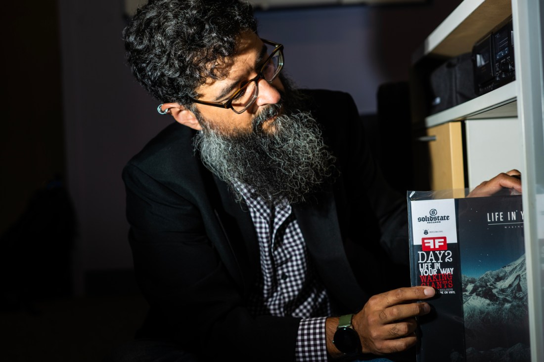 Andrew Mall wearing a black checkered shirt and black blazer looking thoughtfully at a shelf of records. He's using one hand to pull out one of the vinyl records in front of the camera.