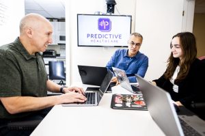 Jeff Berman, Chief Commercial Officer, Talal Ali Ahmad, CEO, and Hailey Koppelman, Client Relationship Associate, sit together at a conference table with laptops during a meeting at Predictive Healthcare in Bedford, Massachusetts. A screen behind them displays the Predictive Healthcare logo.