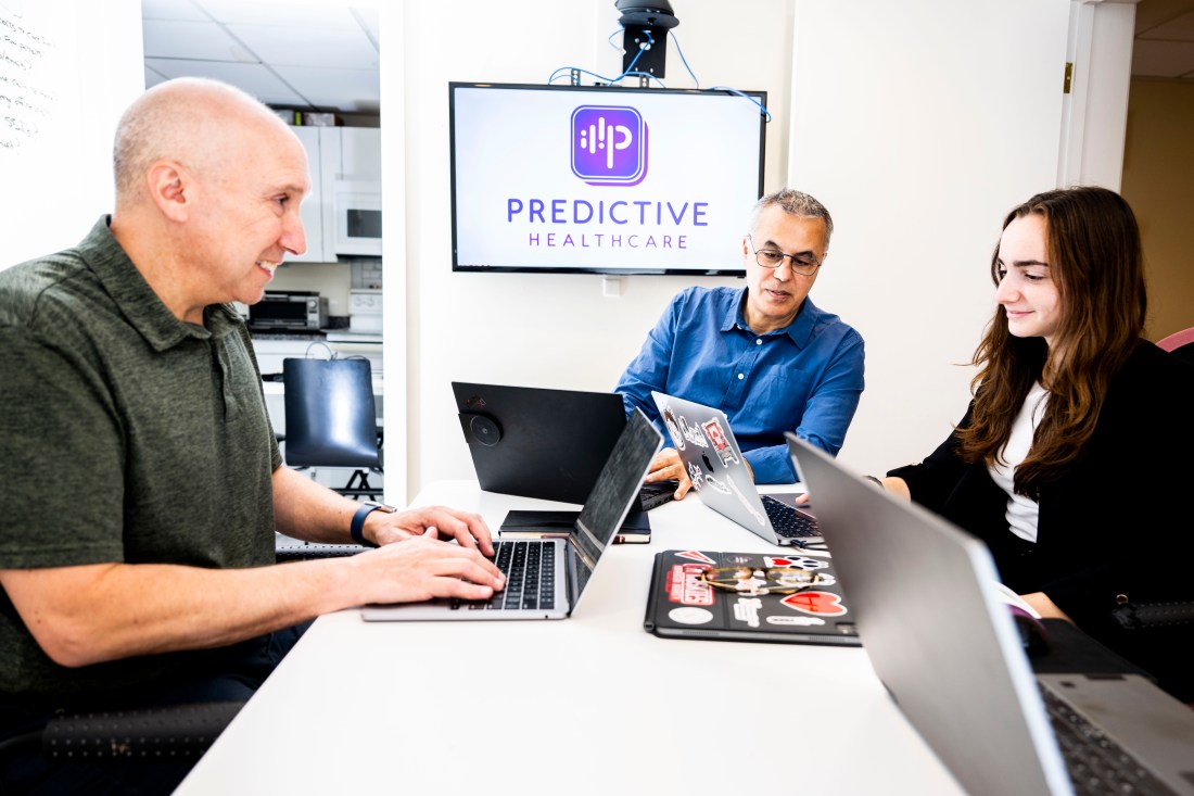 Three team members at Predictive Healthcare—Chief Commercial Officer Jeff Berman, CEO Talal Ali Ahmad, and Client Relationship Associate Hailey Koppelman—collaborate on laptops in a bright office. A monitor behind them displays the Predictive Healthcare logo.