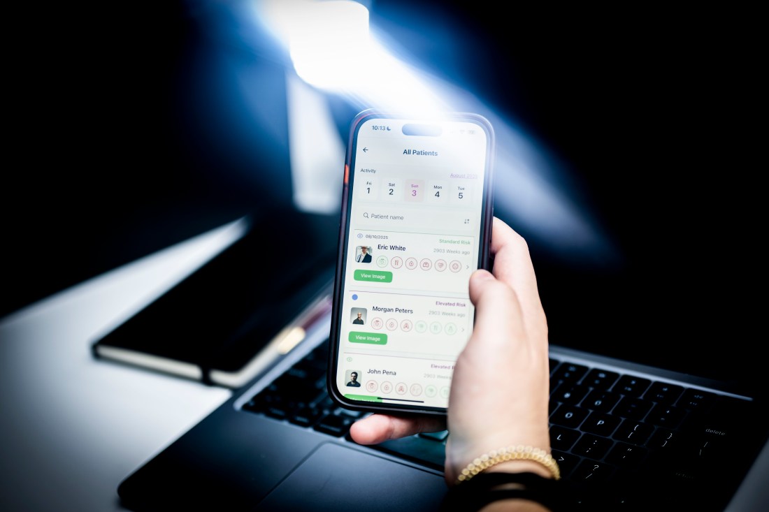 A hand holds a smartphone showing a medical app with a patient list and risk levels, in front of a laptop on a desk.