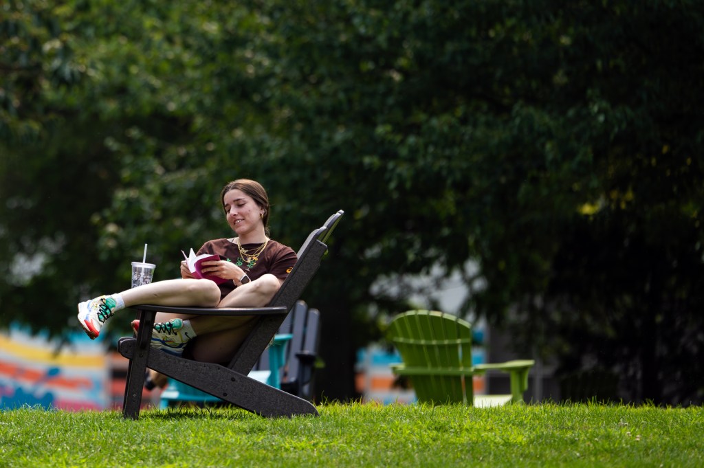 A person sits outside on a lounge chair with their legs on an armrest while reading a novel.
