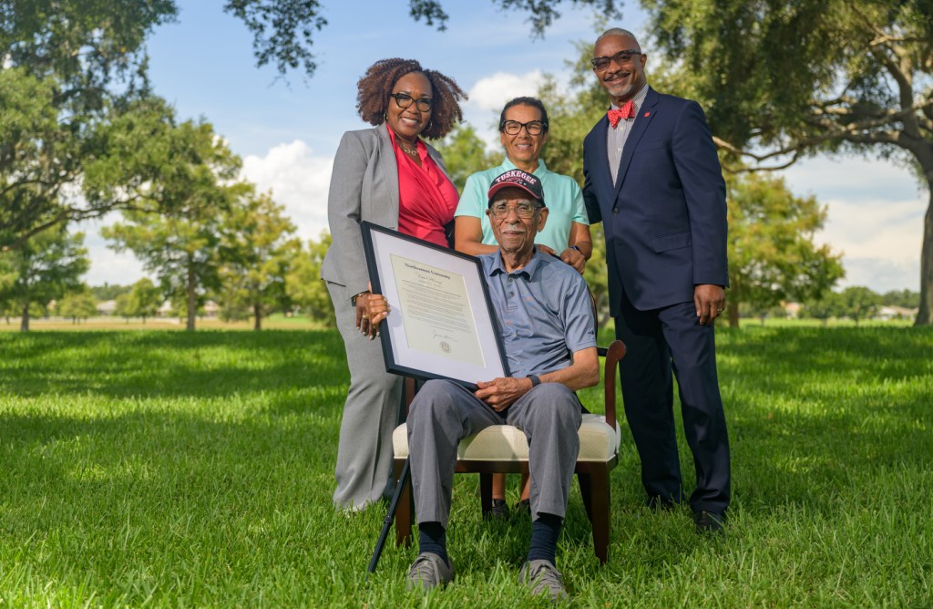 Tuskegee Airman Daniel Keel holds a framed citation while seated on a lawn, joined by his daughter and two presenters.