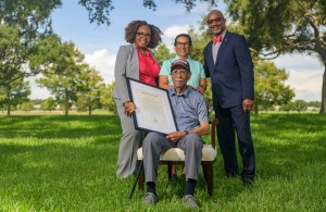 Tuskegee Airman Daniel Keel holds a framed citation while seated on a lawn, joined by his daughter and two presenters.