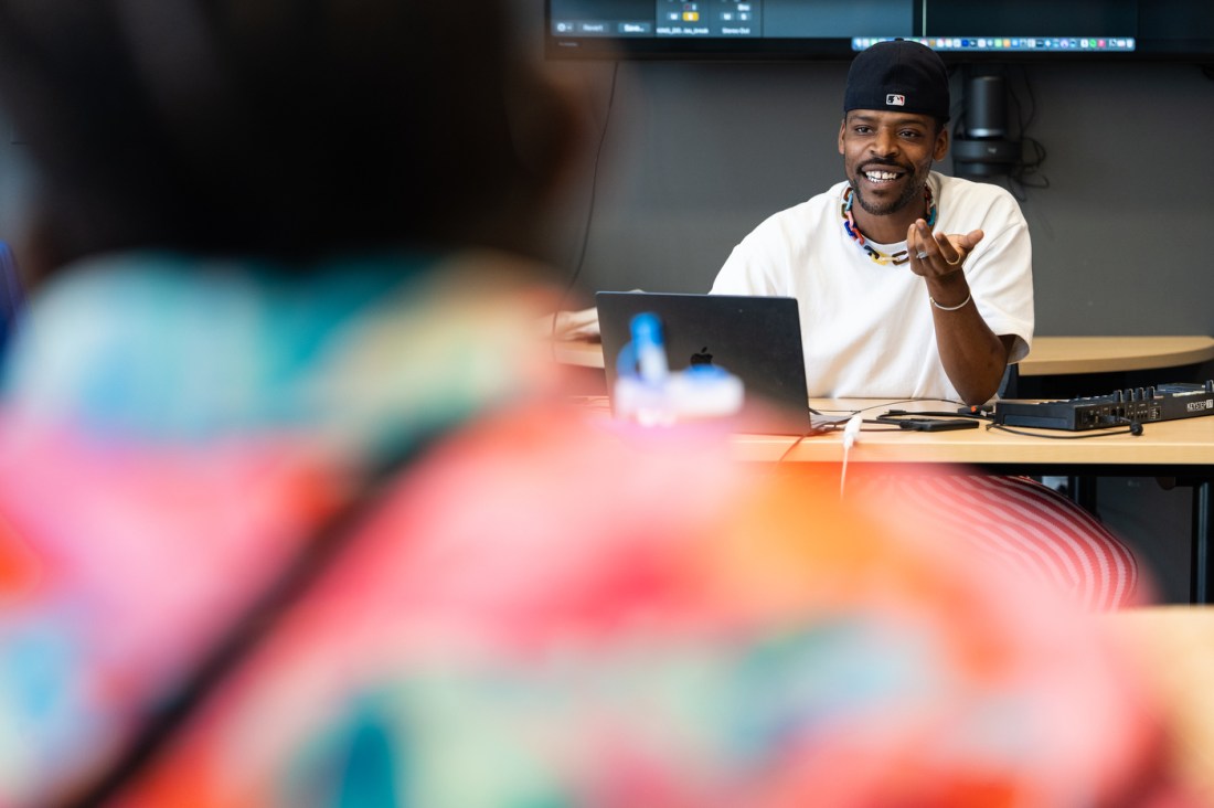 Gogo instructing the room wearing a colorful necklace and ball cap. In the foreground you see the colorful background of a student's hoodie. 