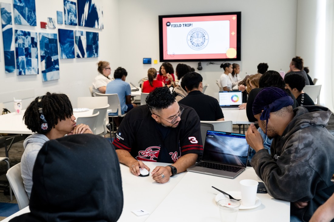 A clean, bright room with a screen on the far wall and a collage. In the foreground students work together clustered at different tables.