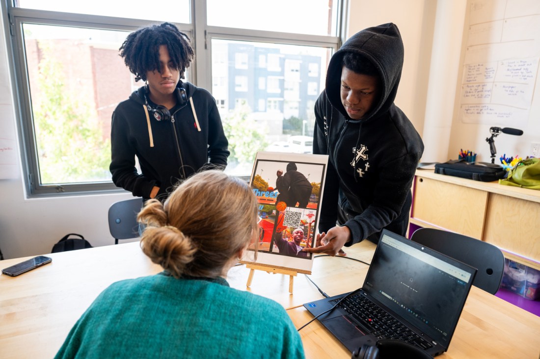 Two students present their work on a tablet to an instructor. One is pointing out a particular detail on the screen of someone raising their arm excitedly.