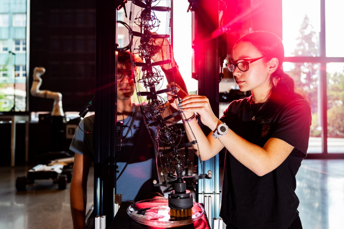 A high school intern adjusts wires on a complex robot while a Northeastern Ph.D. student observes in a bright, modern robotics lab.