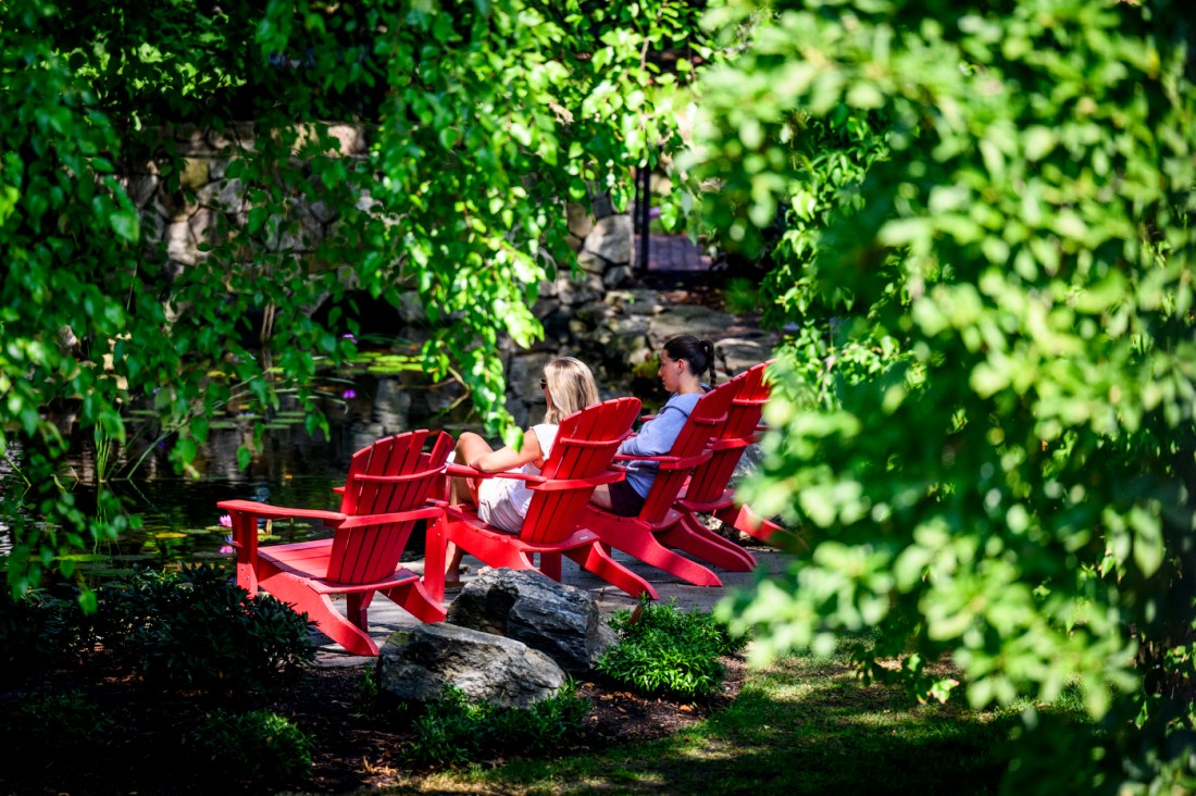 Two people sit in bright red Adirondack chairs beside Northeastern’s koi pond, surrounded by dense green trees and summer foliage.