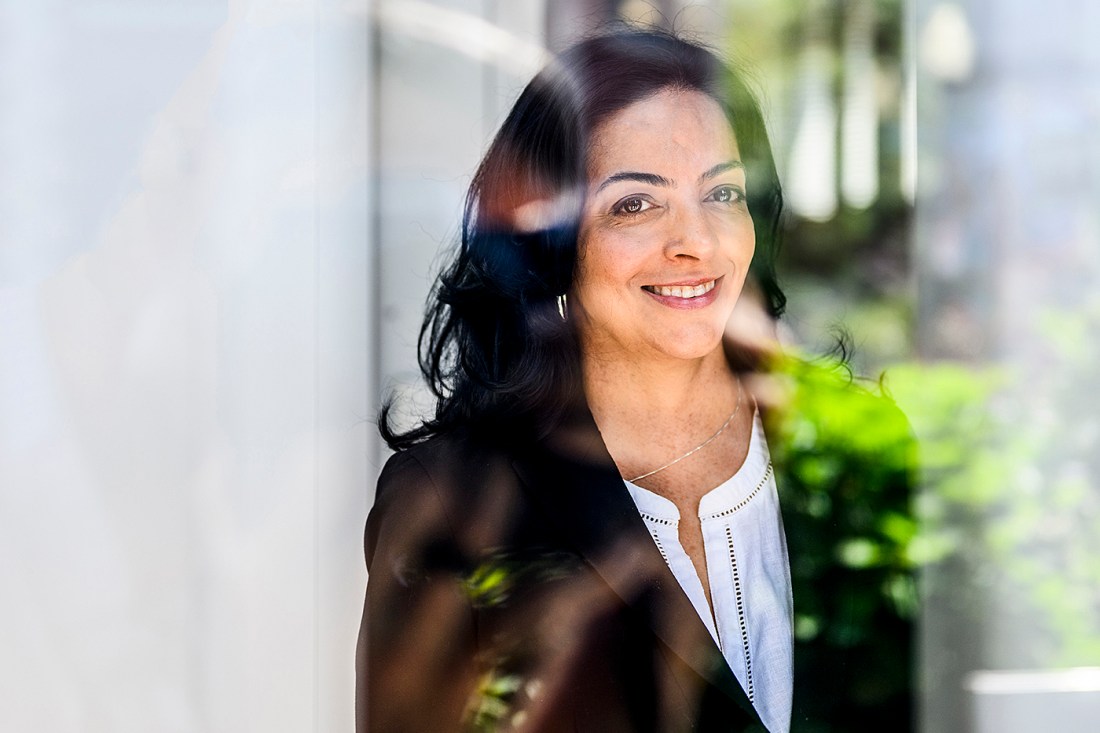 A woman with dark hair and a warm smile stands behind a glass window, wearing a black blazer over a white blouse. Natural light filters through the glass, reflecting greenery and soft shapes from the outside environment.
