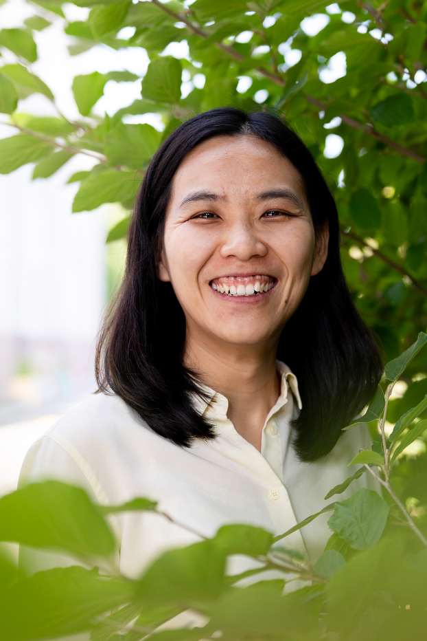 A woman smiles warmly while standing outdoors among green leaves, wearing a cream-colored button-down shirt. Sunlight filters through the foliage, creating a soft, natural background.