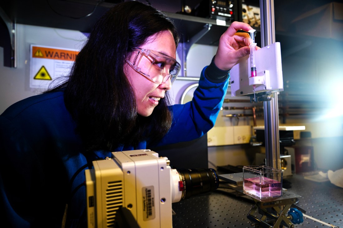 Northeastern professor Xiaoyu Tang conducts research on liquid droplets in the Egan Research Center lab, carefully adjusting a syringe mounted on experimental equipment while wearing protective goggles and a blue lab coat.
