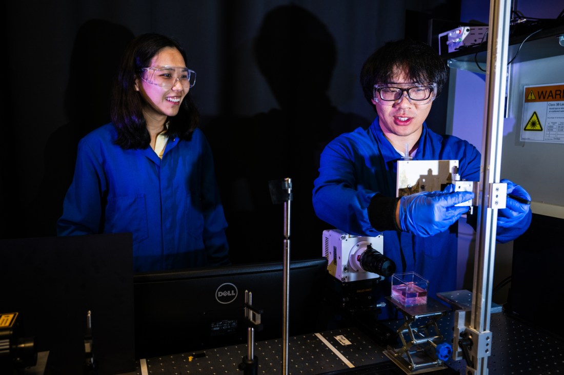 Northeastern professor Xiaoyu Tang and mechanical engineering student Boqian Yan conduct research on liquid droplets using scientific instruments in a lab at the Egan Research Center. Both wear safety glasses and blue lab coats.