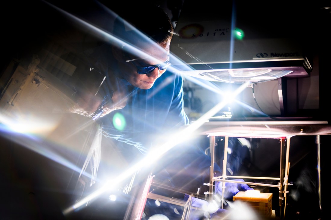 Yi Zheng, wearing safety glasses, examines a solar energy experiment under bright, refracted light beams in a lab at Northeastern University, using high-intensity equipment to study nanoscale materials for sustainable energy solutions.