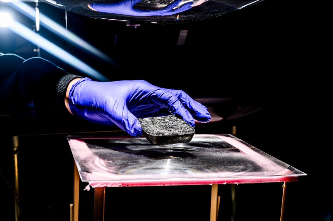A researcher wearing bright blue gloves carefully handles a sample beneath a light source in a lab setting, studying nanoscale materials for solar energy applications at Northeastern University’s Mugar Life Sciences Building.