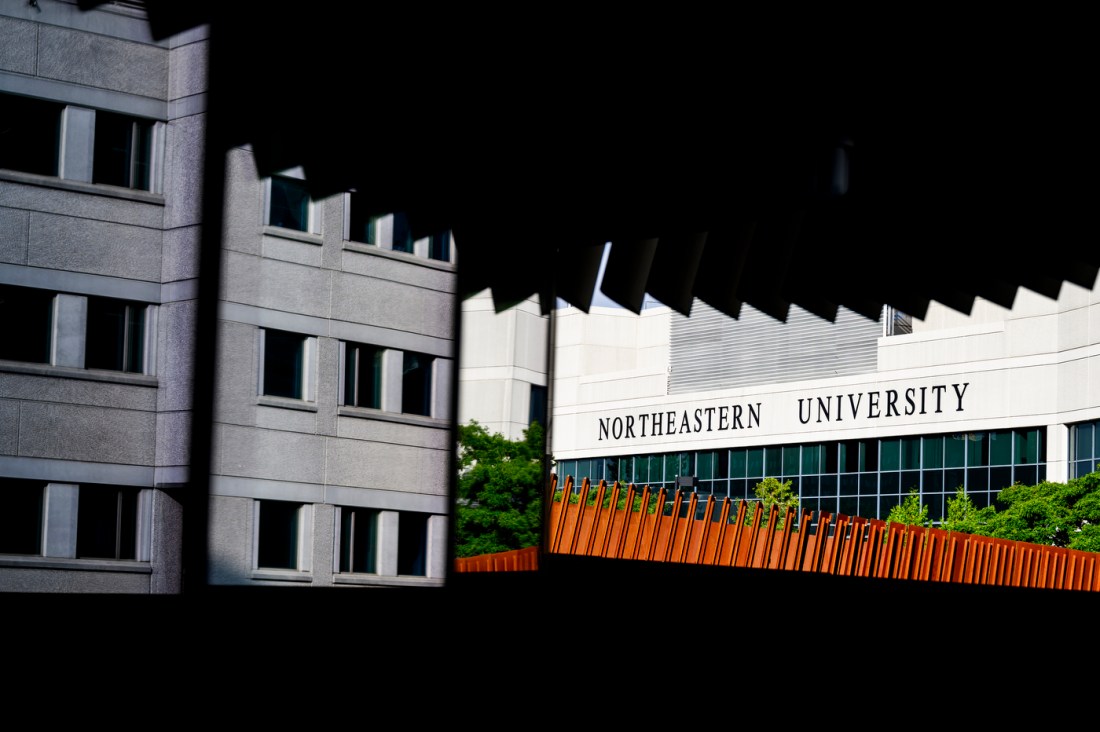 Architectural view of Northeastern University's Boston campus, partially framed by dramatic shadows and angles from an overhead structure. The white building with 'NORTHEASTERN UNIVERSITY' signage stands behind a bright orange fence and green trees, captured on a sunny day.