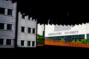 Architectural view of Northeastern University's Boston campus, partially framed by dramatic shadows and angles from an overhead structure. The white building with 'NORTHEASTERN UNIVERSITY' signage stands behind a bright orange fence and green trees, captured on a sunny day.