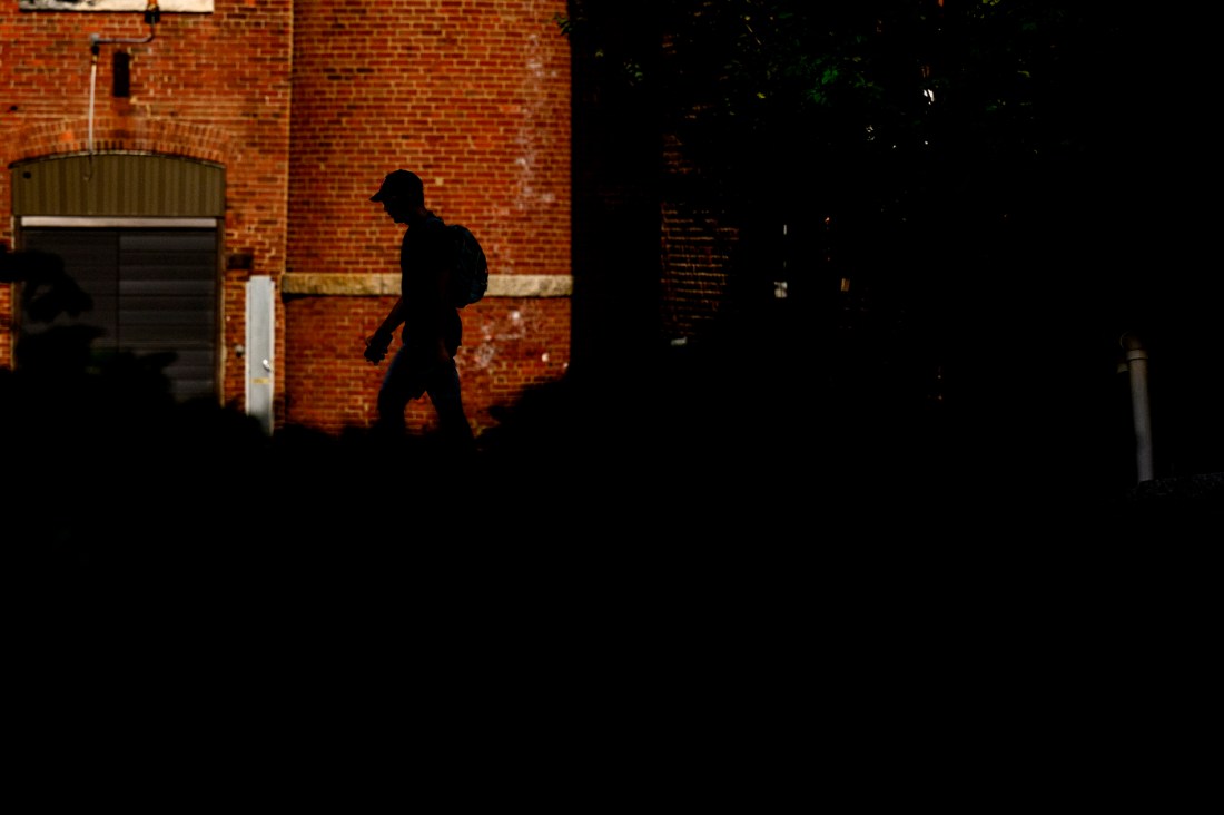 A student with a backpack walks past a brick building on Northeastern University’s Boston campus, silhouetted in shadow against the sunlit wall. The contrast between light and dark highlights the architectural texture and campus atmosphere.