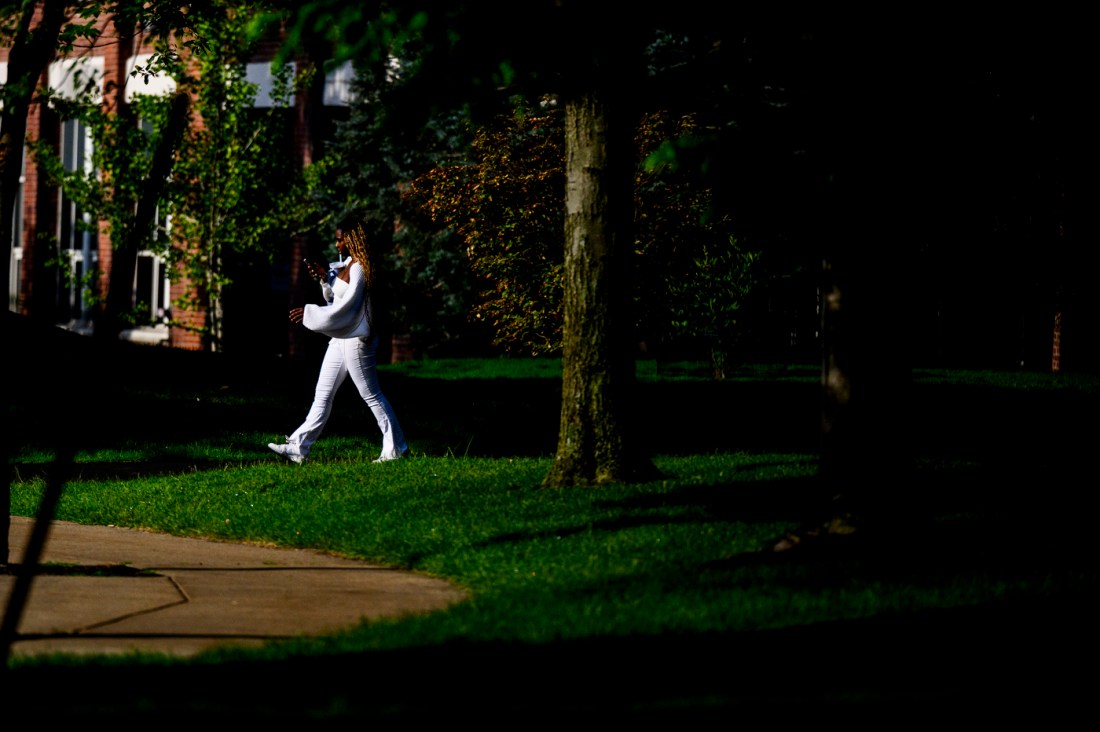 A student in white clothing walks through Northeastern University’s Boston campus, surrounded by lush green trees and red-brick buildings in the background. The scene captures a peaceful, shaded moment on campus in late summer.