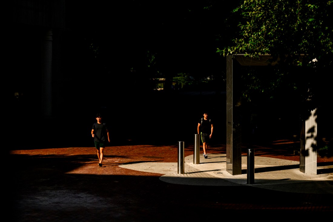 Two students walk through a sunlit path on Northeastern University’s Boston campus, framed by dramatic shadows and architectural structures. The contrast between the bright walkway and the surrounding darkness emphasizes the golden hour lighting.