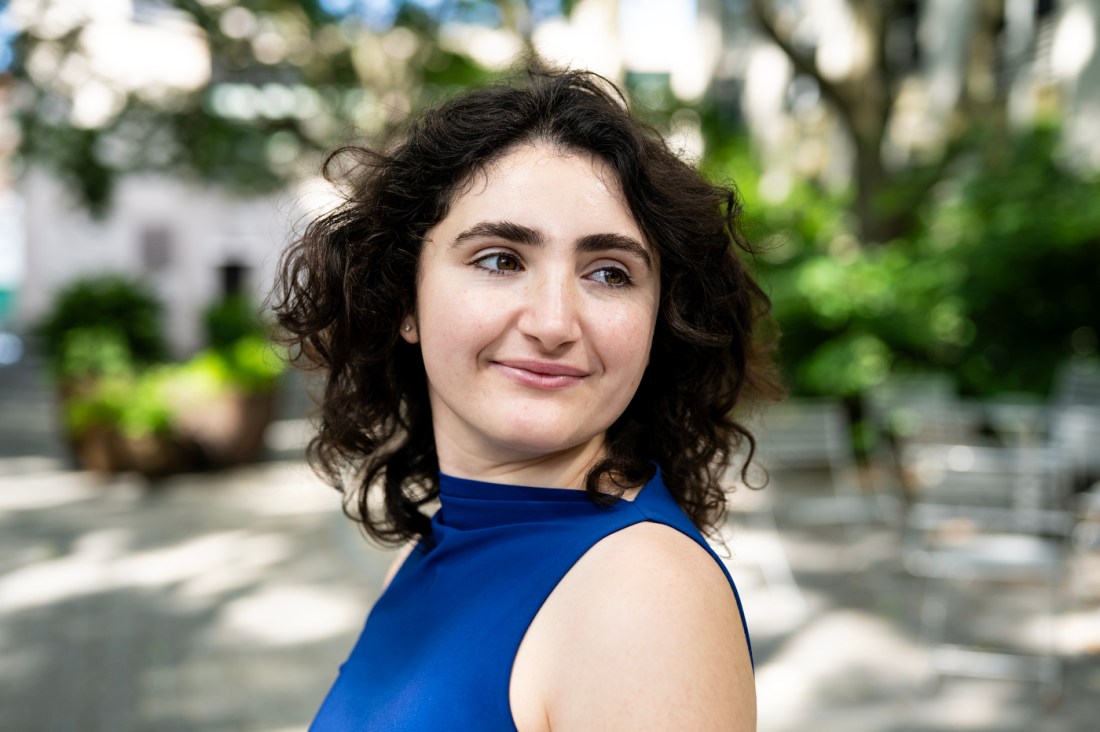 A woman with curly dark hair and a sleeveless royal blue top smiles softly while glancing over her shoulder in an outdoor setting filled with greenery and blurred café-style chairs.