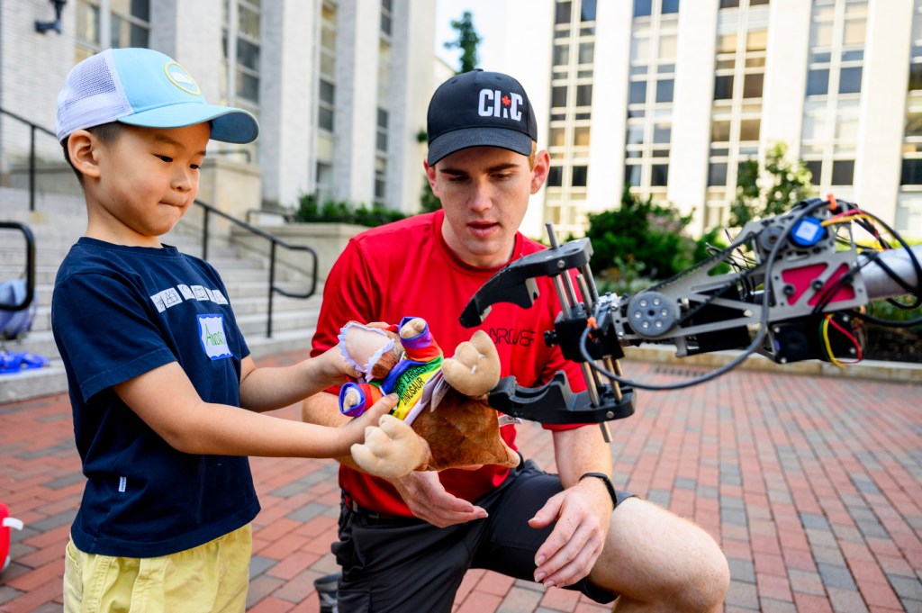 A mechanical engineering student shows a Mars Rover model to young children from the R.J. Call Children’s Center at an outdoor university quad.