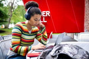 A student wearing headphones sits outside while working on a laptop.