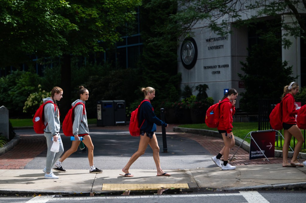 Six Northeastern University women’s soccer players walk in a line along a shaded campus sidewalk, wearing athletic gear and red backpacks, heading to Parsons Field for a game.